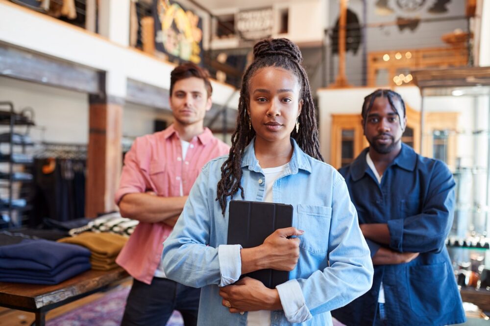 Three retail workers standing in a clothing shop, the woman in front holding a tablet